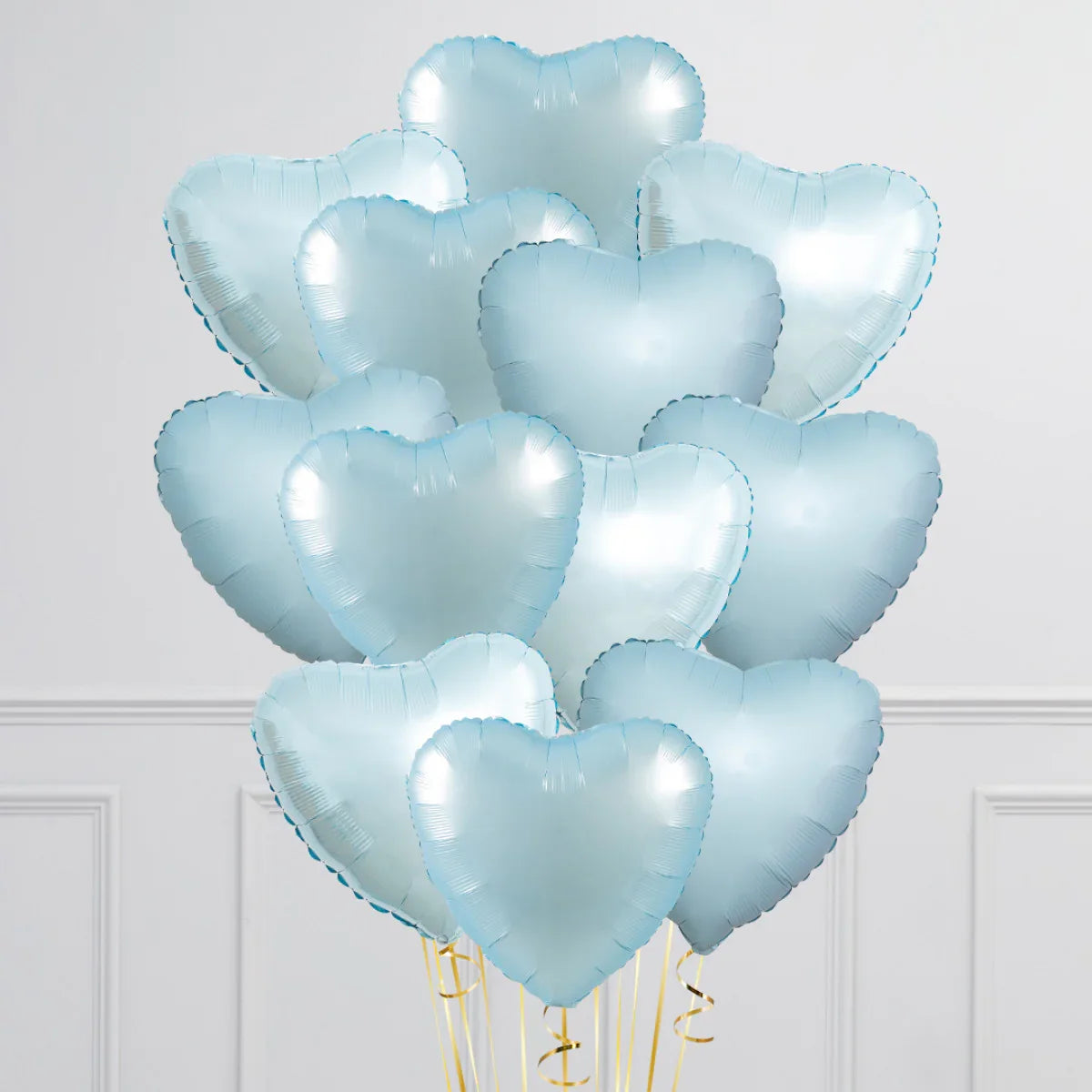 Bunch of shiny baby blue heart-shaped foil balloons with golden ribbons against a white wall