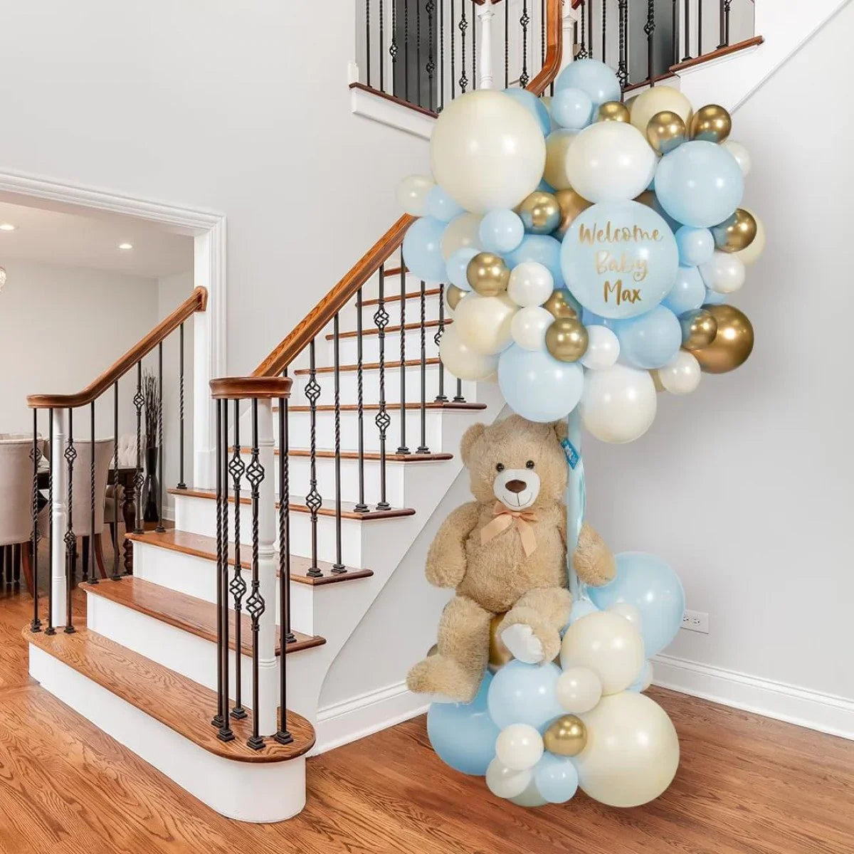Baby shower decoration with blue and gold balloons, teddy bear, and 'Welcome Baby Max' sign by stairs