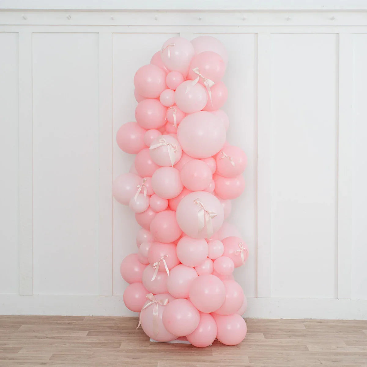 Tall pink balloon pillar decorated with light pink balloons and white ribbon bows against white wall