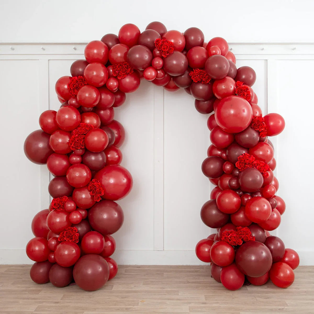 Dark cherry red balloon arch with clusters of red flowers indoors on wooden floor
