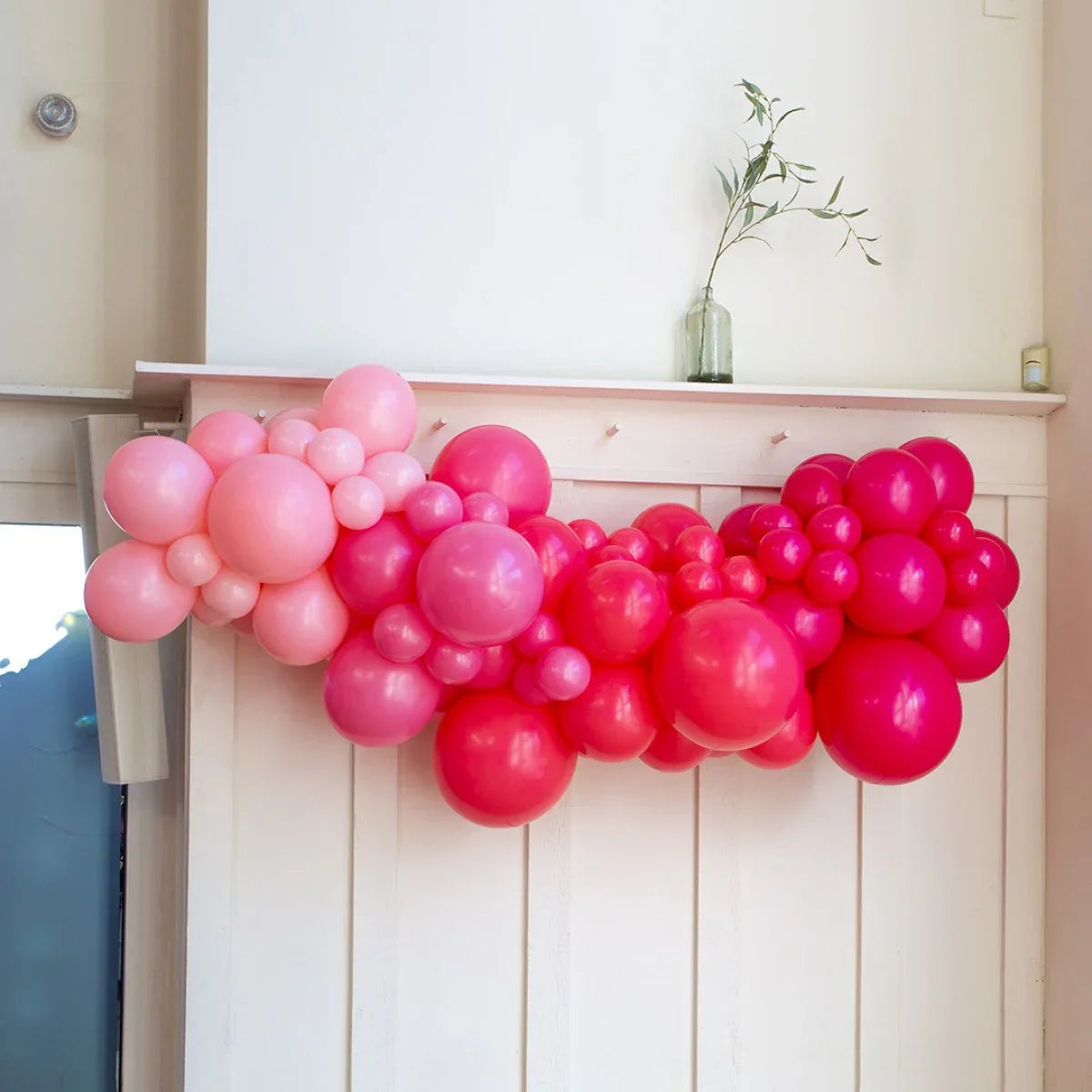 Pink and red balloon garland decoration above white wooden paneling with green vase and candle