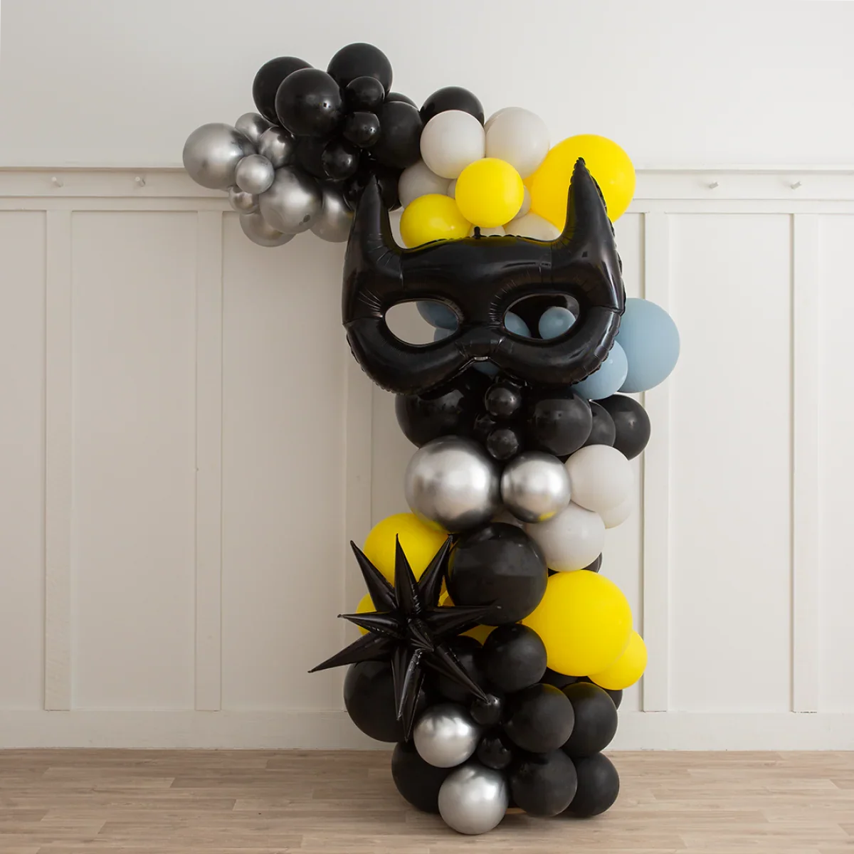 Asymmetric balloon arch in black, yellow, silver, gray, and blue with a large black Batman mask balloon and star-shaped balloon