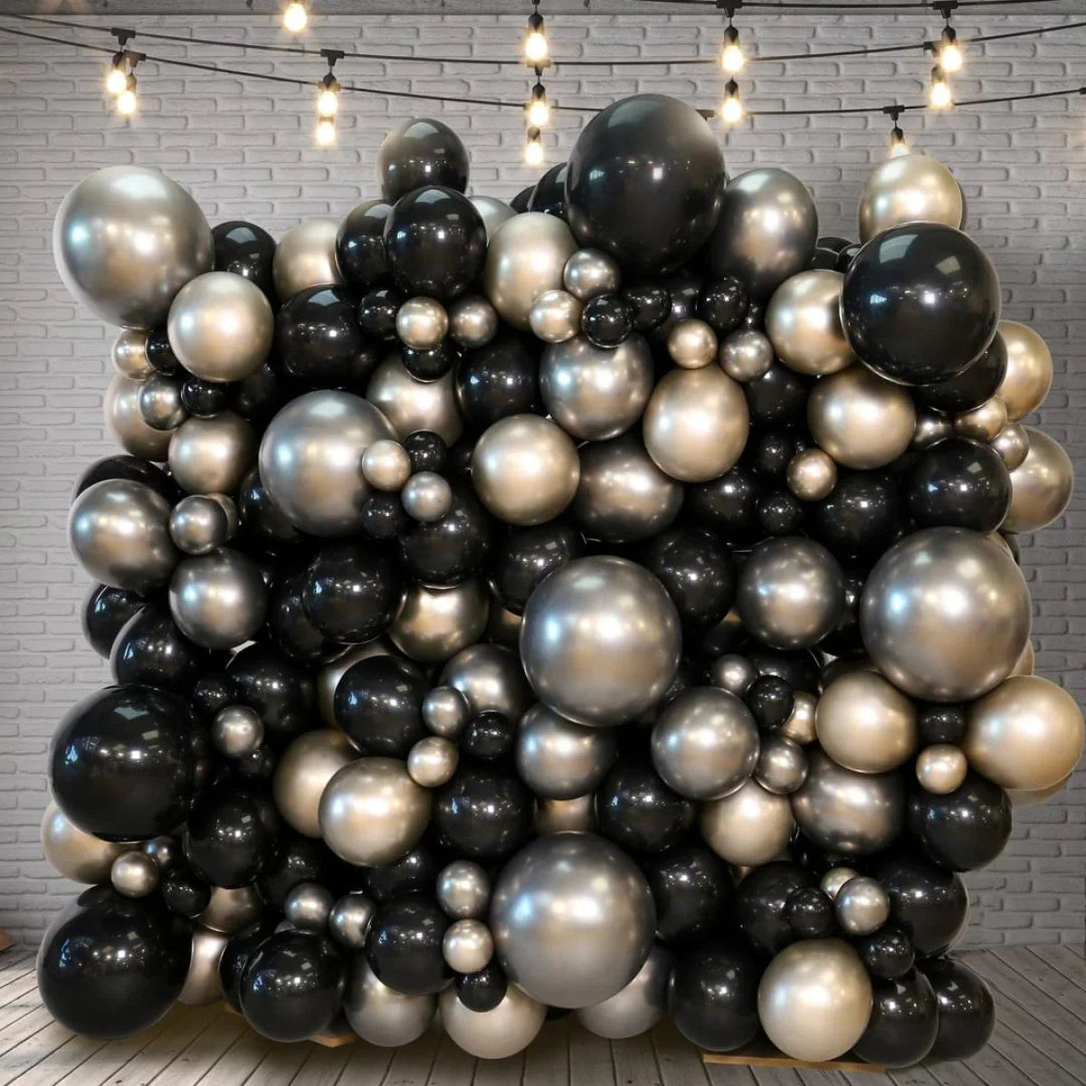 Decorative balloon wall in black and champagne gold against gray brick backdrop with string lights overhead