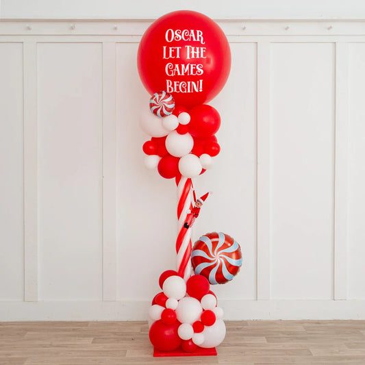 Red and white balloon column with peppermint decorations and elf toy, giant red balloon says 'Oscar Let The Games Begin'