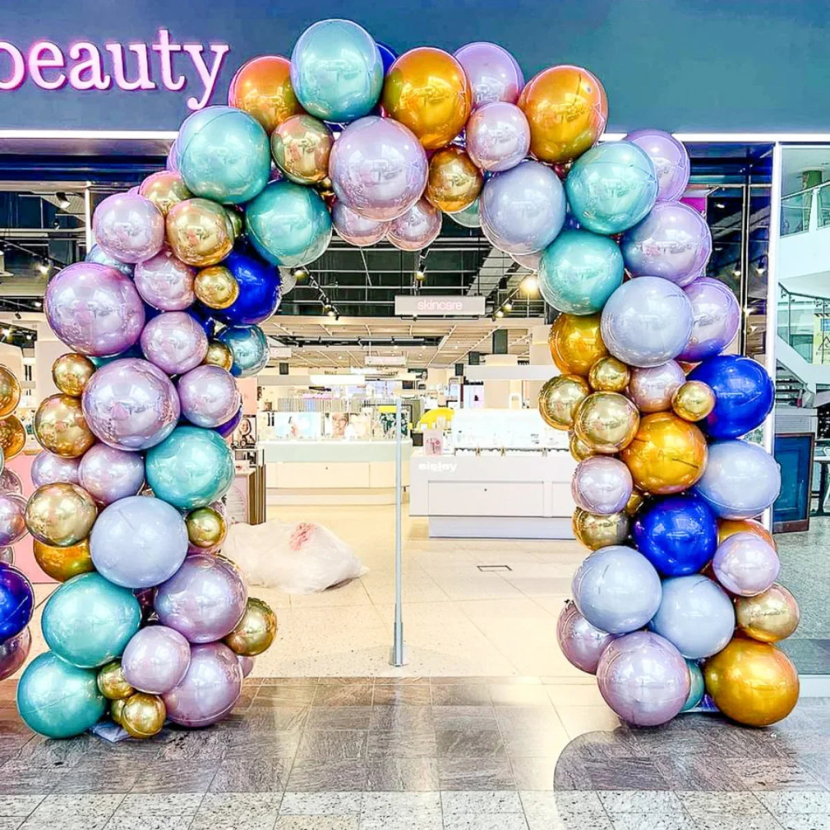 Colorful balloon arch with metallic gold, blue, lavender, and teal balloons at a beauty store entrance inside a mall