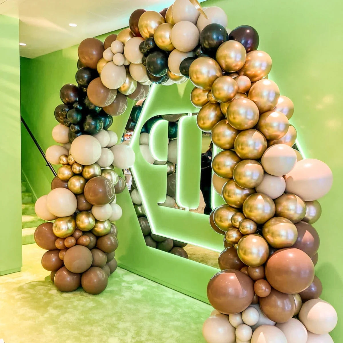 Luxurious balloon arch in gold, brown, beige with glowing green geometric frame at indoor event