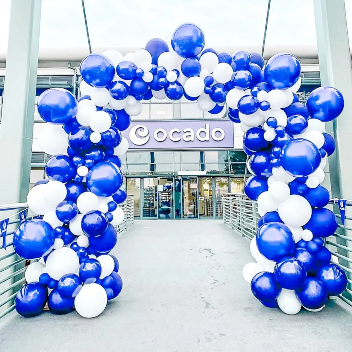 Blue and white balloon arch at entrance of Ocado building with glass doors and railings
