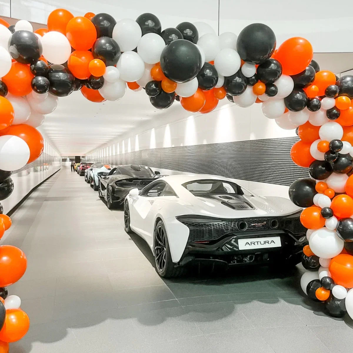 Luxury McLaren Artura and sports cars lined up under orange, black, and white balloon arch in modern showroom