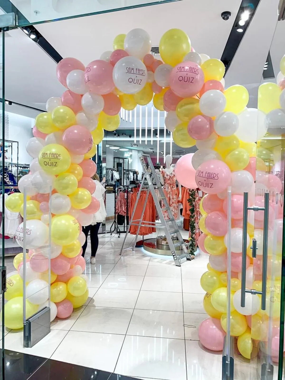 Colorful balloon arch in pastel pink, white, and yellow at a retail store entrance with clothes and ladder inside