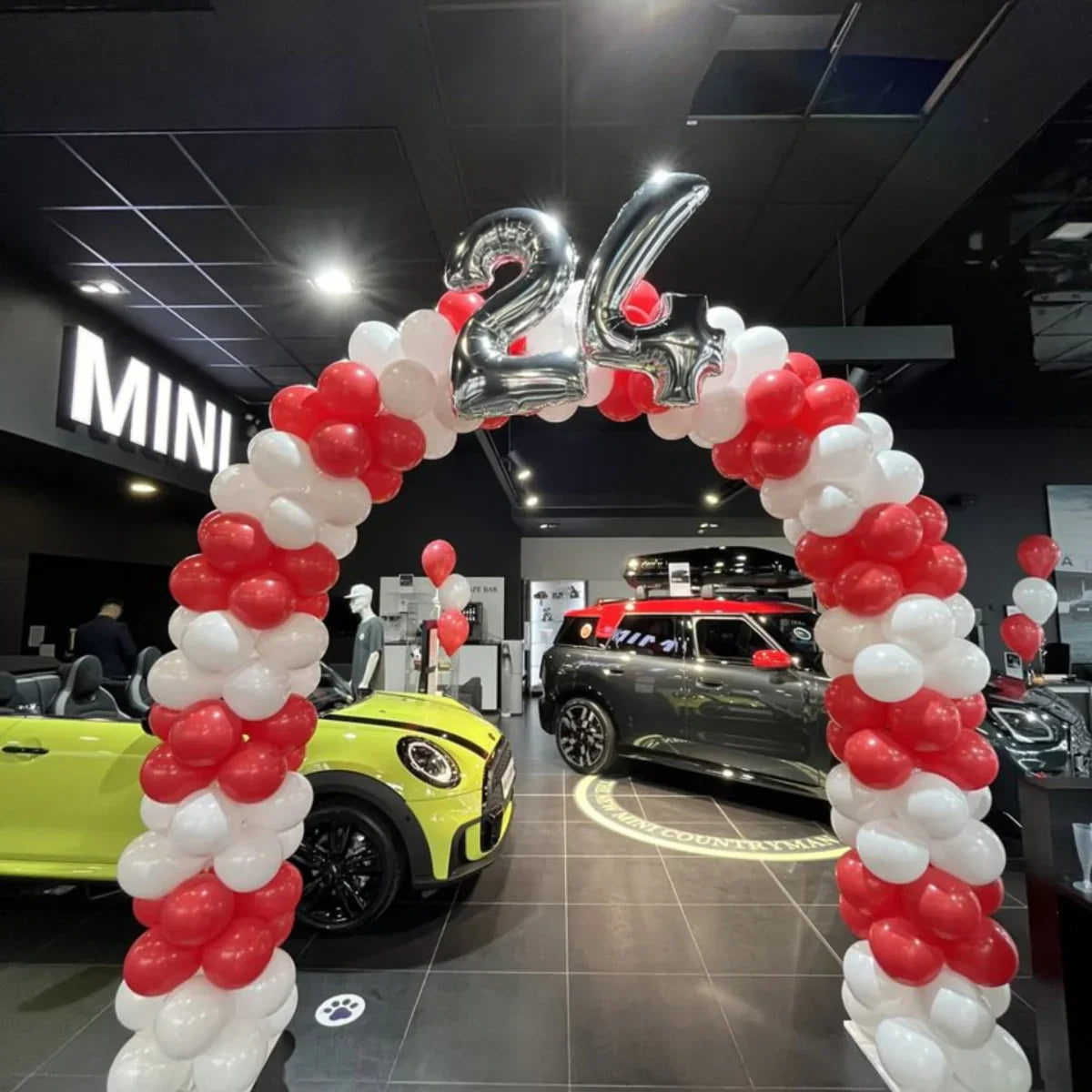 Red and white balloon arch with silver 24 balloons at a MINI car showroom featuring yellow and black MINI cars