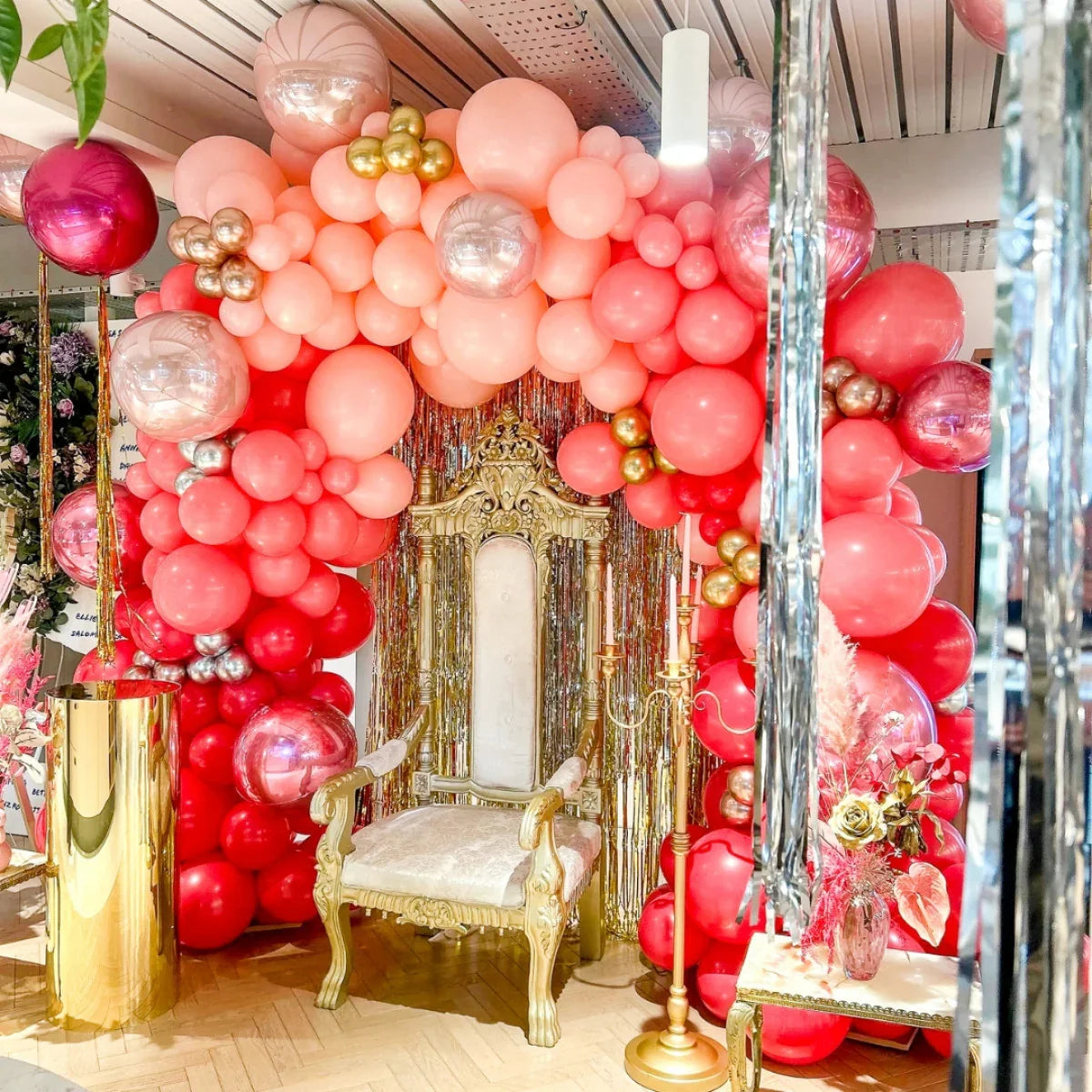 Elegant gold throne chair under pink and red balloon arch with metallic accents in event decor