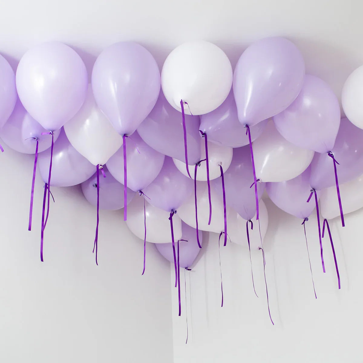 Lavender and white helium balloons with purple ribbons clustered on a white ceiling corner