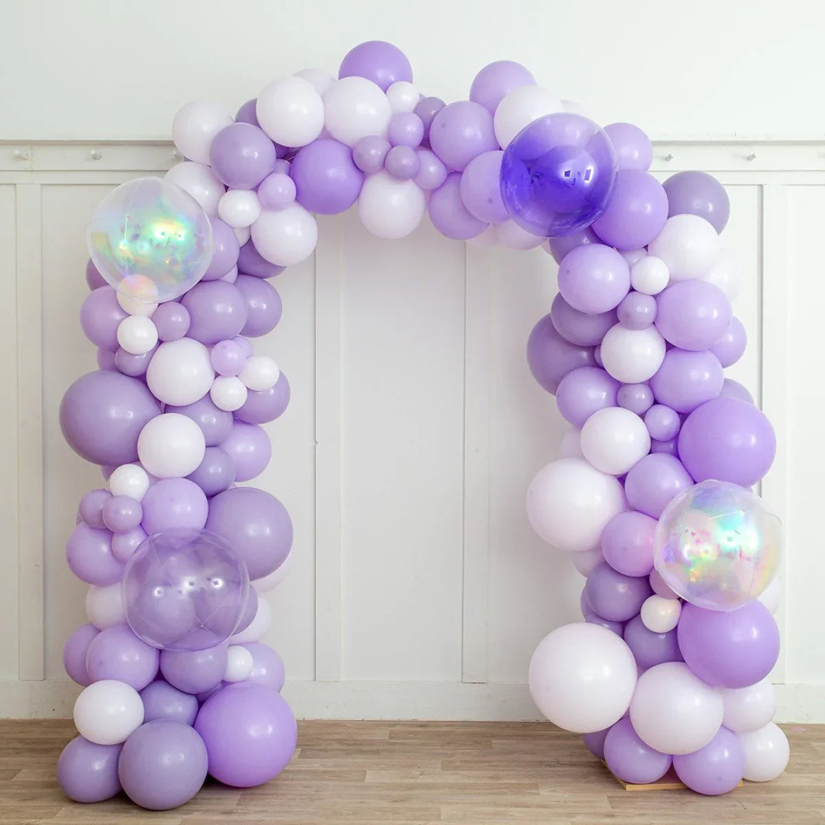 Lavender and white balloon arch with iridescent and purple balloons on wooden floor and white wall
