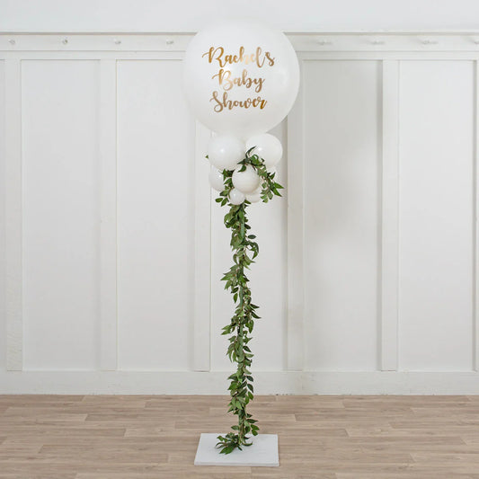 White balloon stand with gold text "Rachel's Baby Shower" and green leaf garland decoration