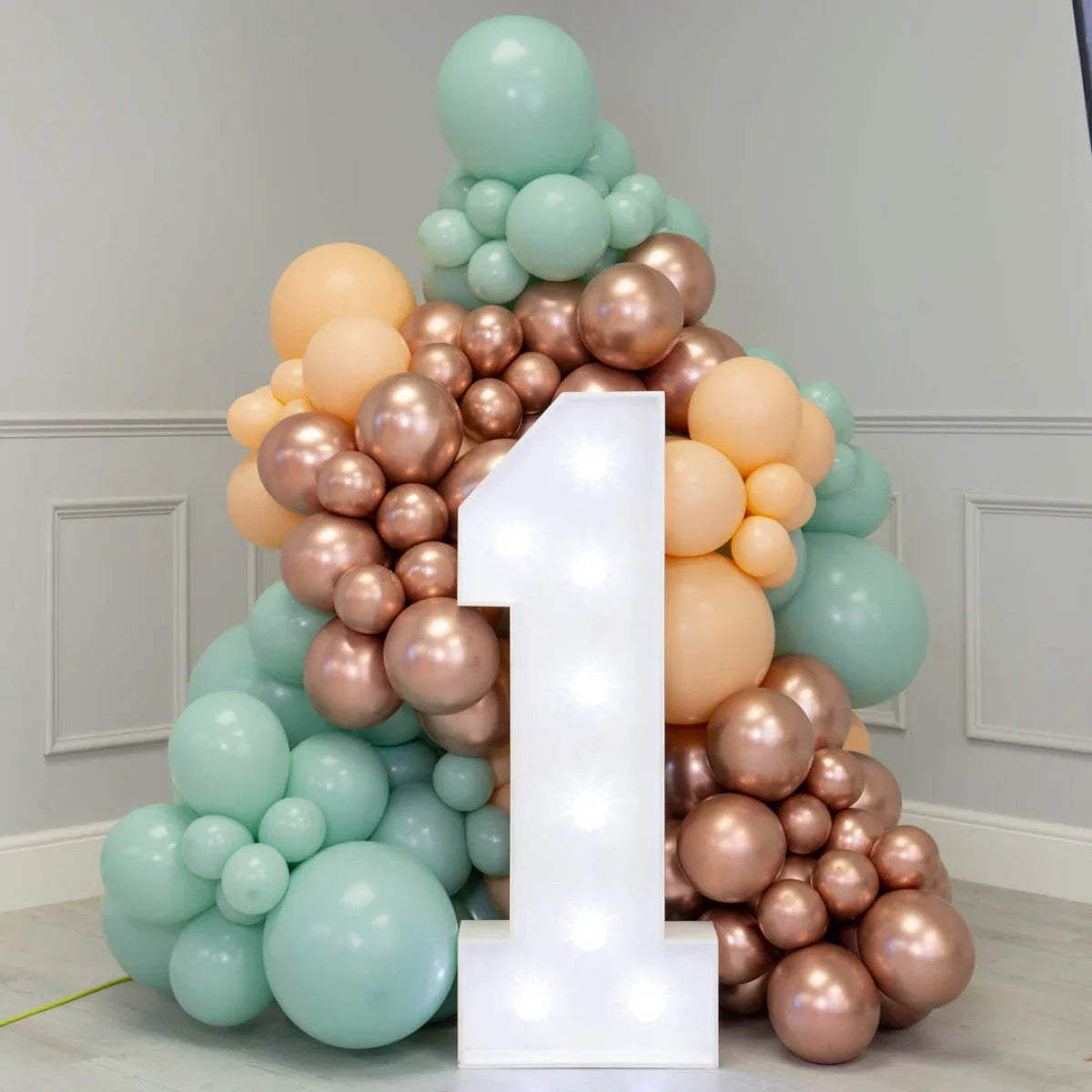 Light-up number 1 sign surrounded by pastel and metallic balloon arrangement in soft green, peach, and bronze colors