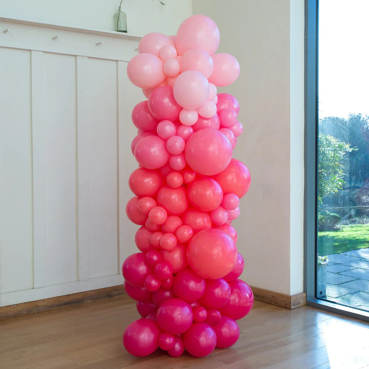 Tall pillar of pink gradient balloons in an indoor room with wooden floor and window