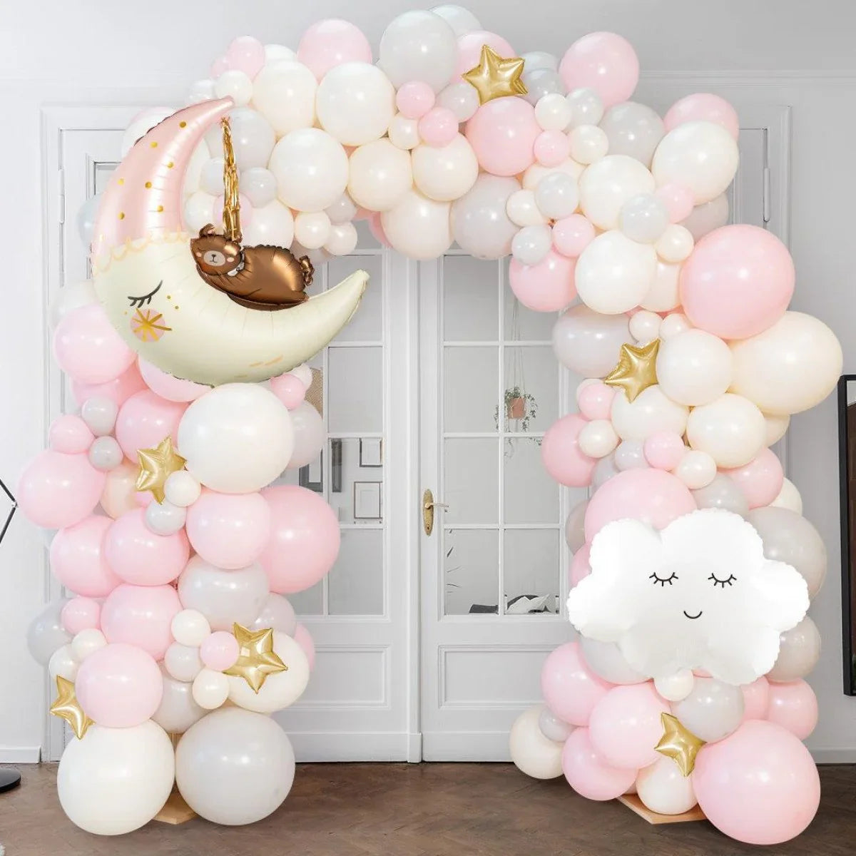 Pink and white balloon arch with gold star balloons, moon holding a teddy bear, and smiling cloud balloon