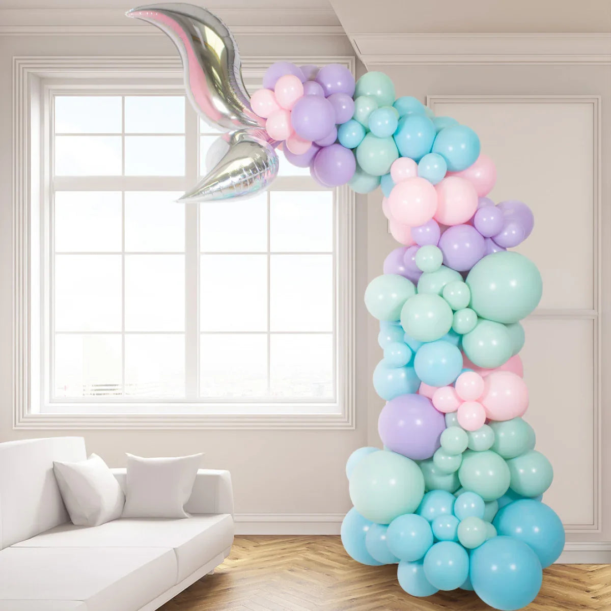 Pastel mermaid tail balloon arch with metallic fin next to white couch in bright room