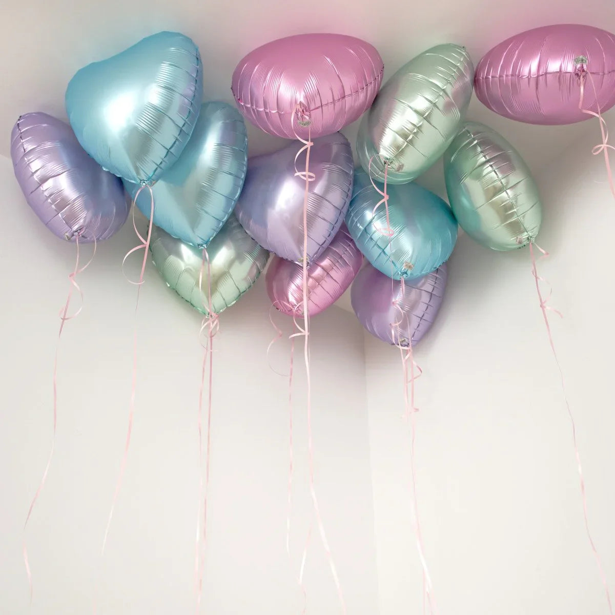 Metallic heart-shaped helium balloons in pastel pink, blue, purple, and silver floating near a white ceiling
