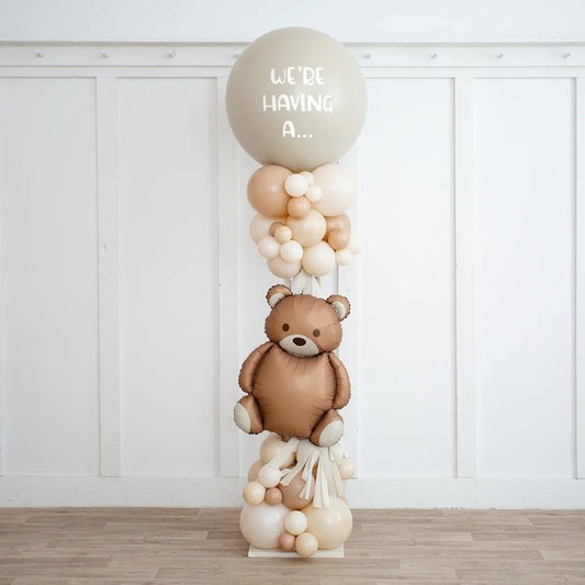 Neutral beige balloon pillar with teddy bear balloon and text we're having a for gender reveal party