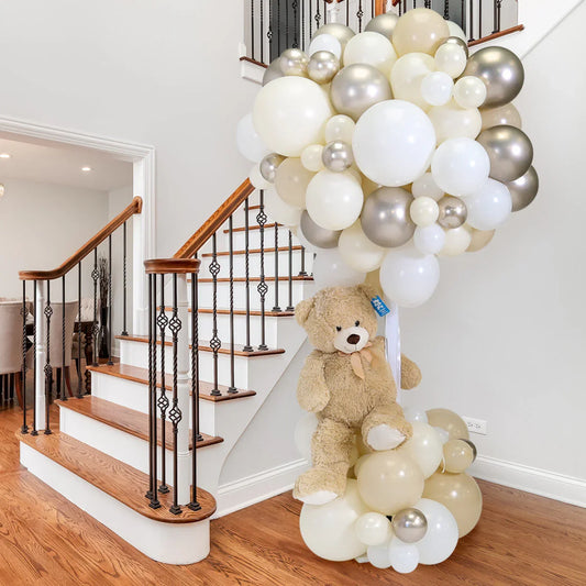 Large teddy bear sitting under a cream and gold balloon arrangement in a modern hallway