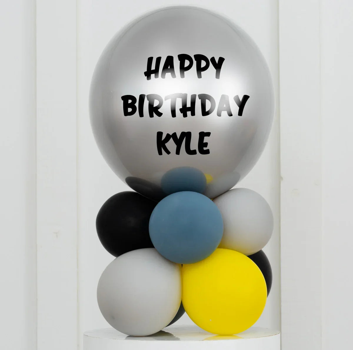 Personalized silver happy birthday Kyle balloon on a stack of black, gray, blue, and yellow balloons