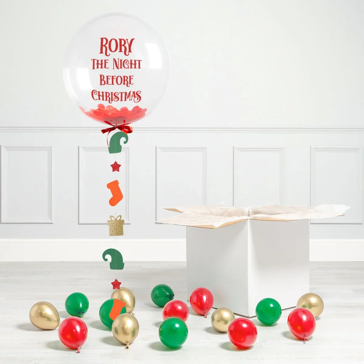 Personalized clear Christmas balloon with red confetti and hanging festive cutouts, surrounded by red, green, and gold balloons