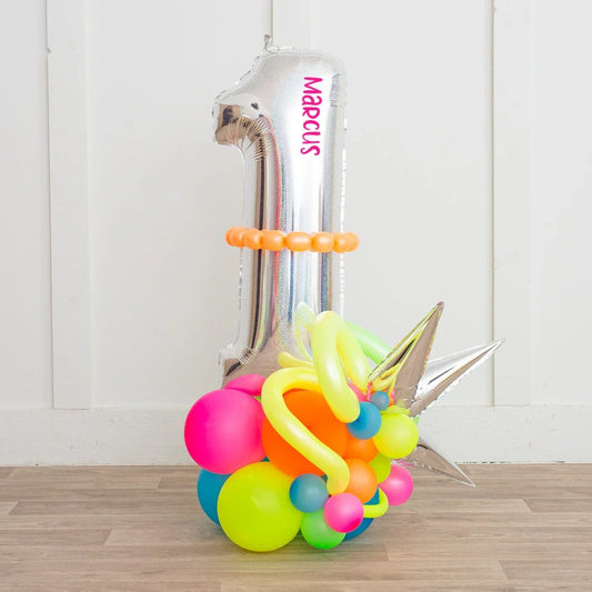 Silver number 1 balloon with personalized text Marcus surrounded by colorful neon balloons on wooden floor