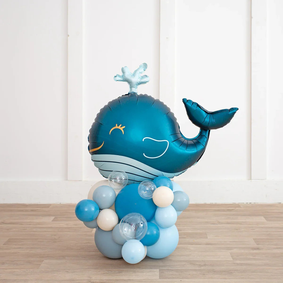 Blue whale-shaped foil balloon with blue, white, and clear latex balloon cluster on wooden floor