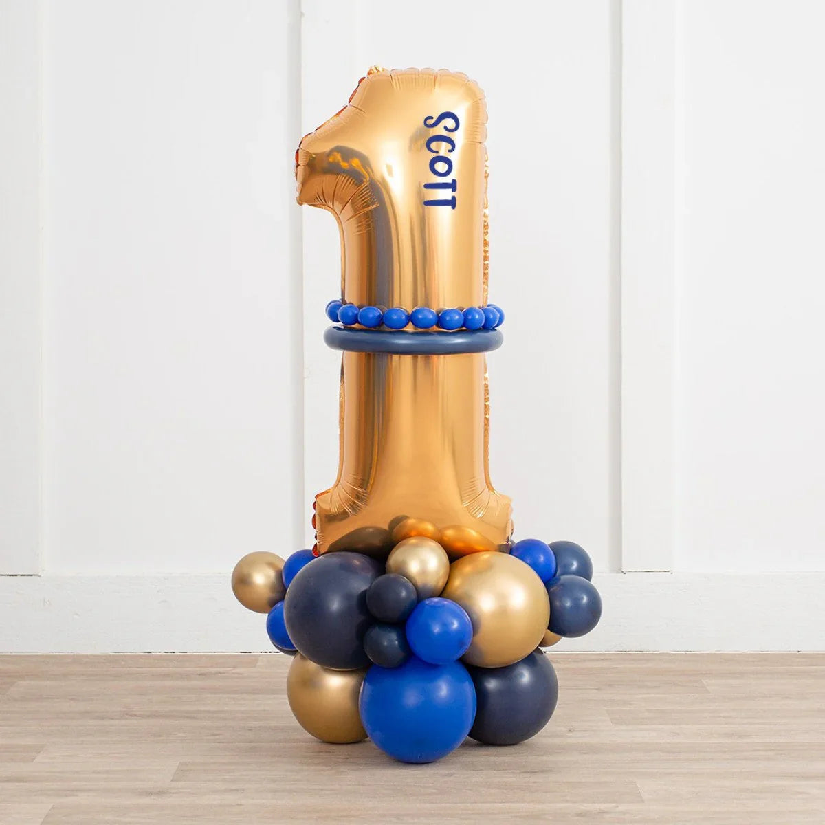 Gold foil number 1 balloon with personalized name Scott surrounded by blue and gold balloons on wooden floor