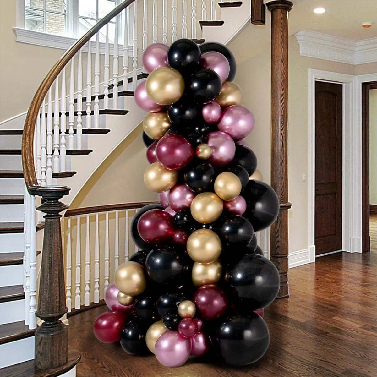 Tall balloon pillar in a home interior with a curved staircase, featuring black, gold, burgundy, and pink balloons