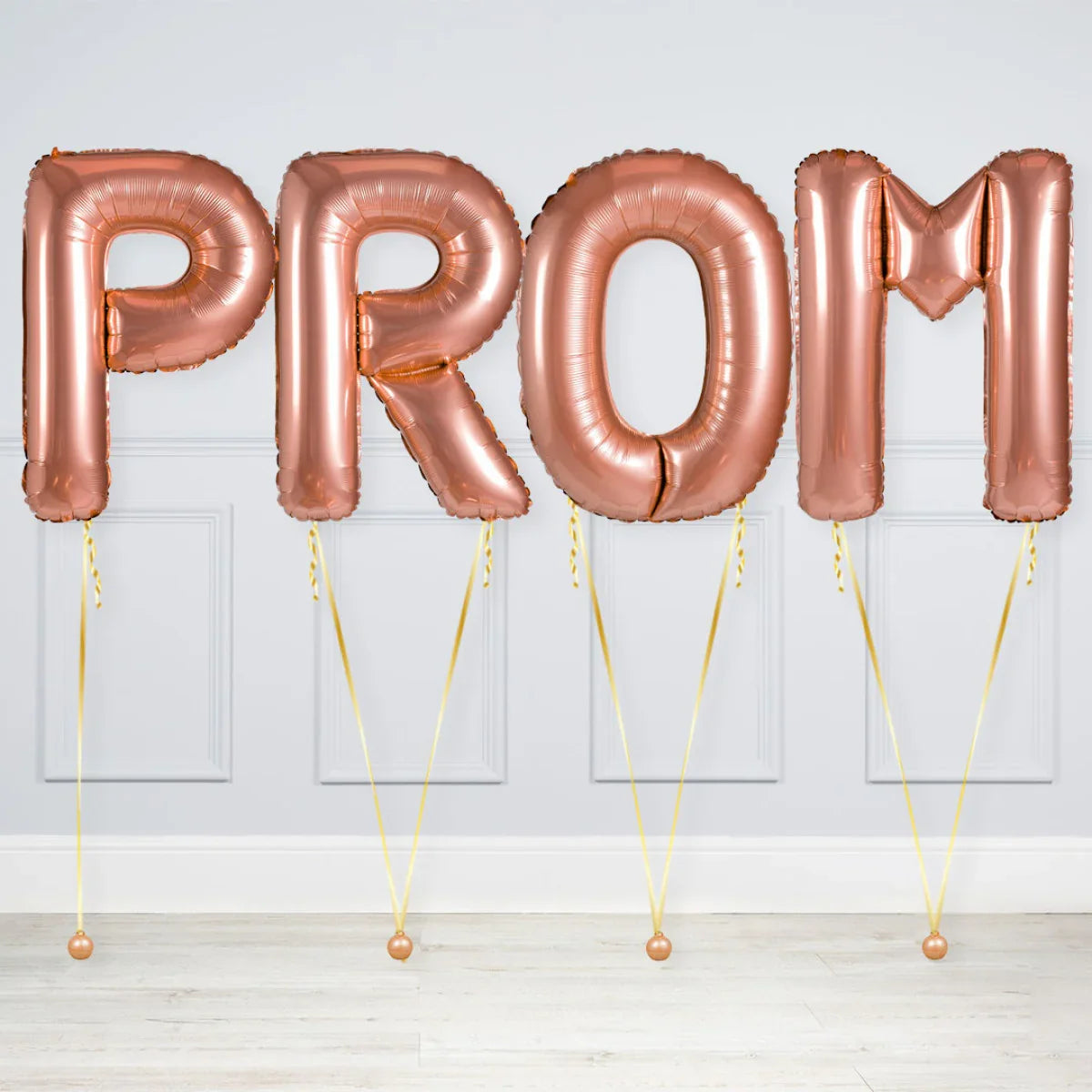 Rose gold foil balloons spelling PROM tied with golden strings against a white wall