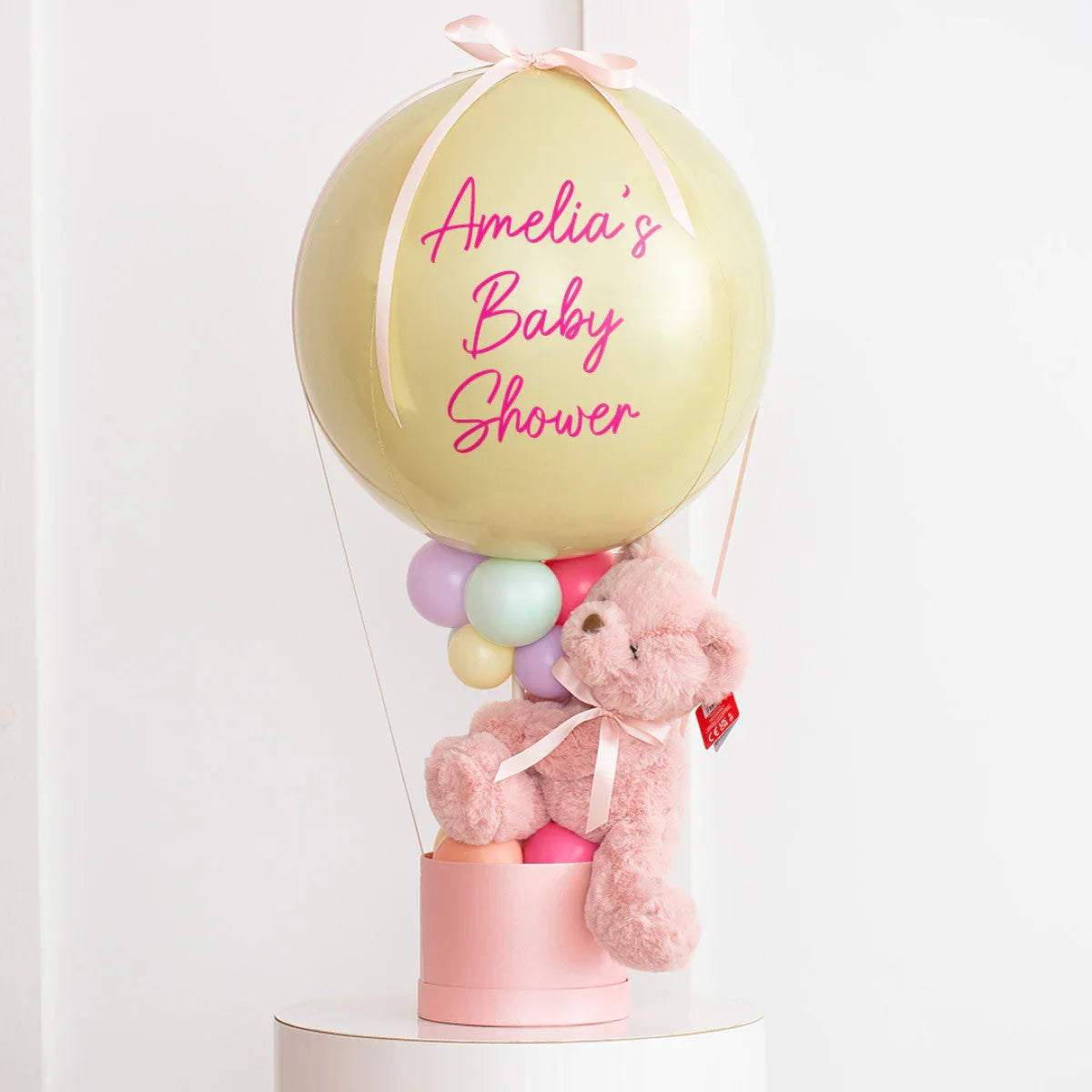 Golden balloon with pink text Amelia's Baby Shower, pastel balloons, and pink teddy bear in pink basket
