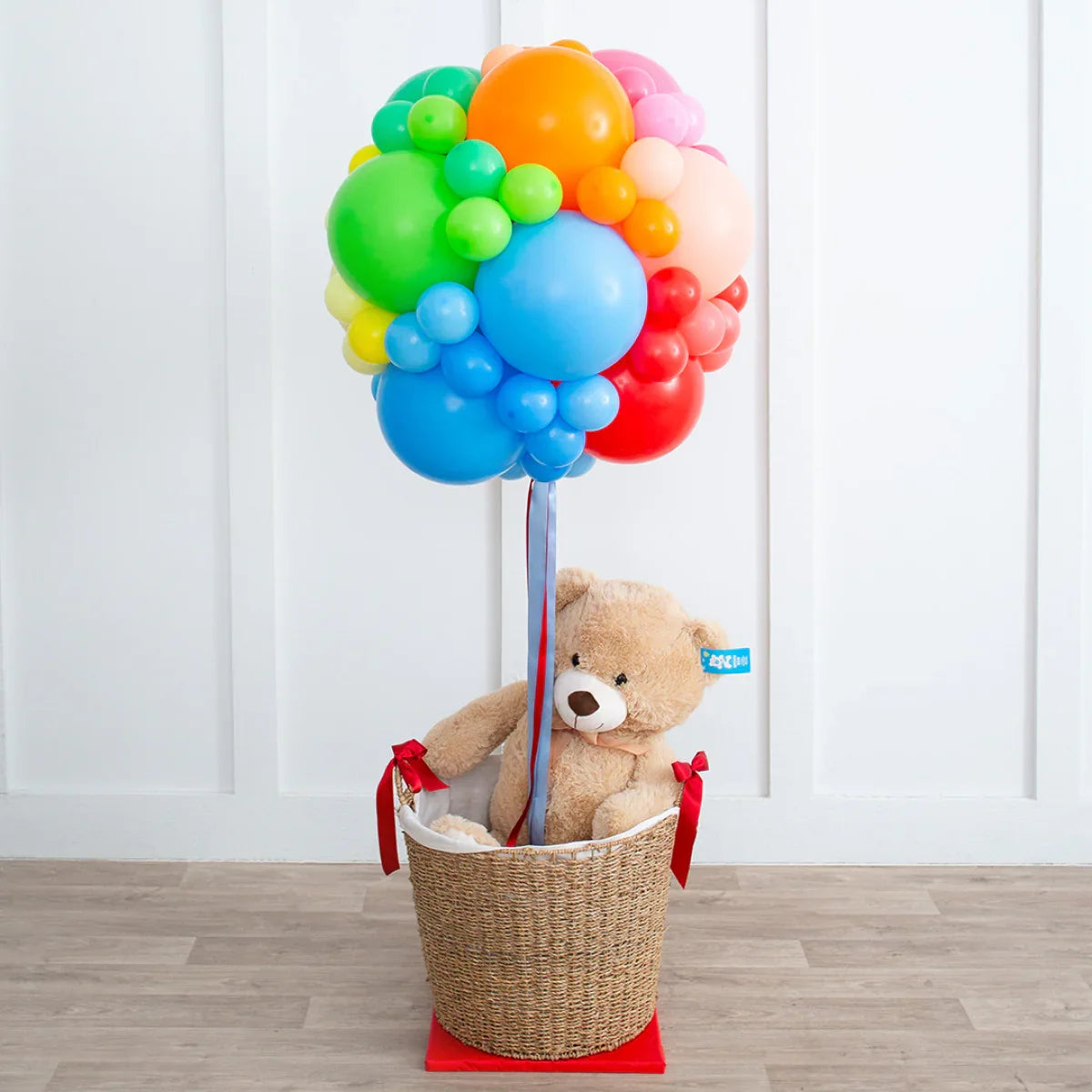 Colorful rainbow balloon bouquet attached to a basket with a plush teddy bear inside on wooden floor