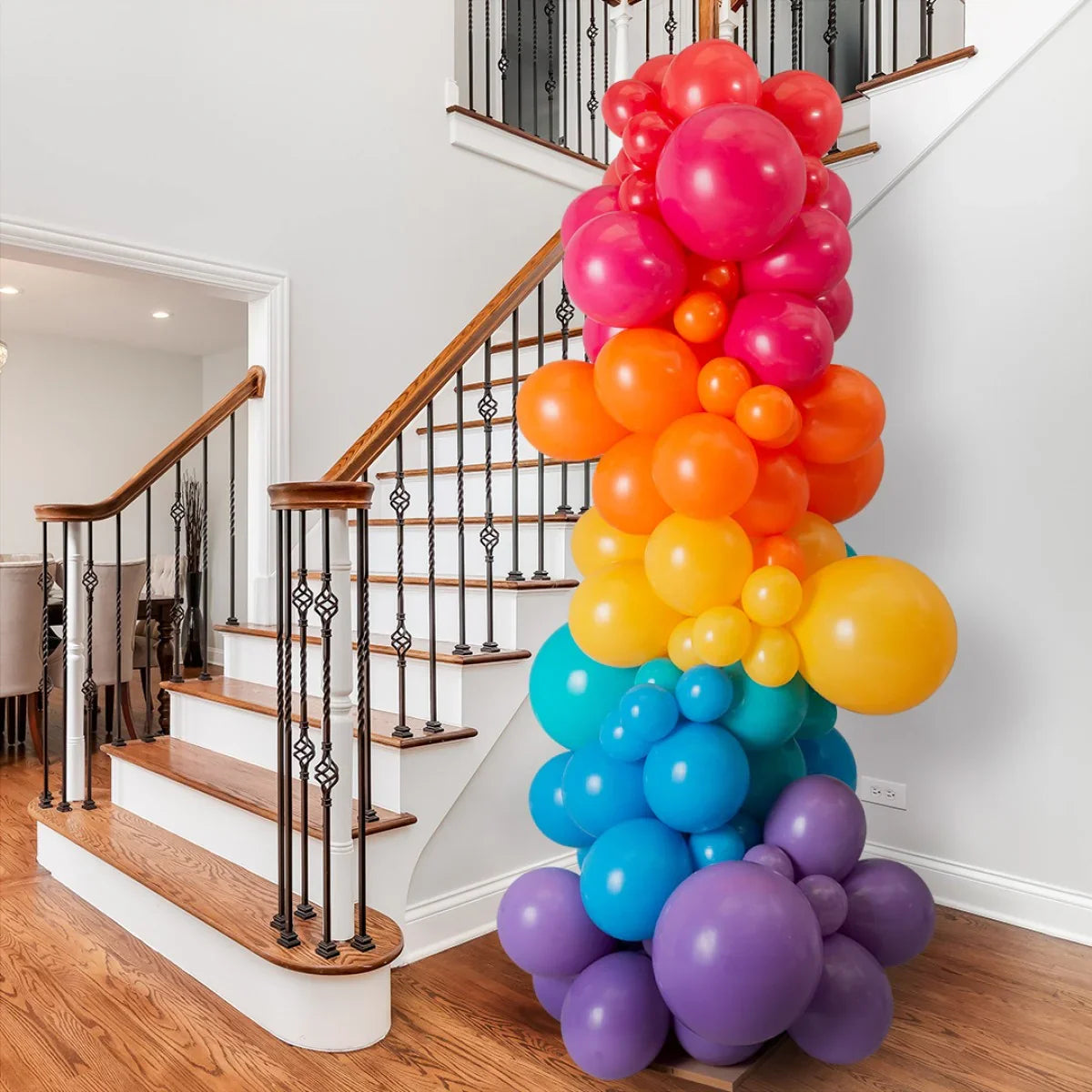 Tall rainbow balloon pillar with purple, blue, yellow, orange, and red balloons beside wooden staircase in modern home