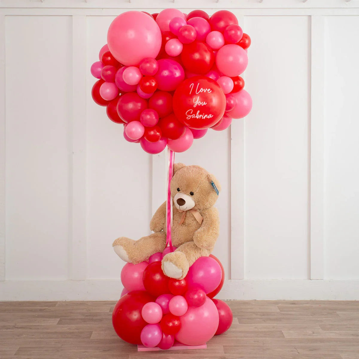 Teddy bear sitting among red and pink balloon clusters with one balloon reading I love you Sabrina