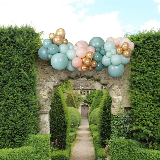 Sage green, blush pink, and gold balloon garland decorating stone archway in lush garden pathway