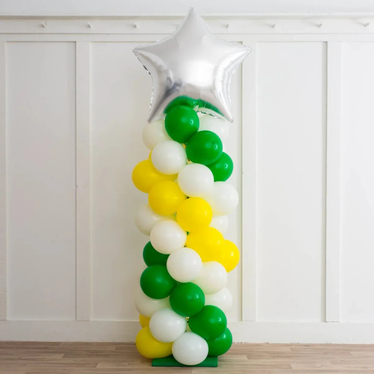 Spiral balloon pillar with green, yellow, white balloons topped by a large silver star in indoor setting