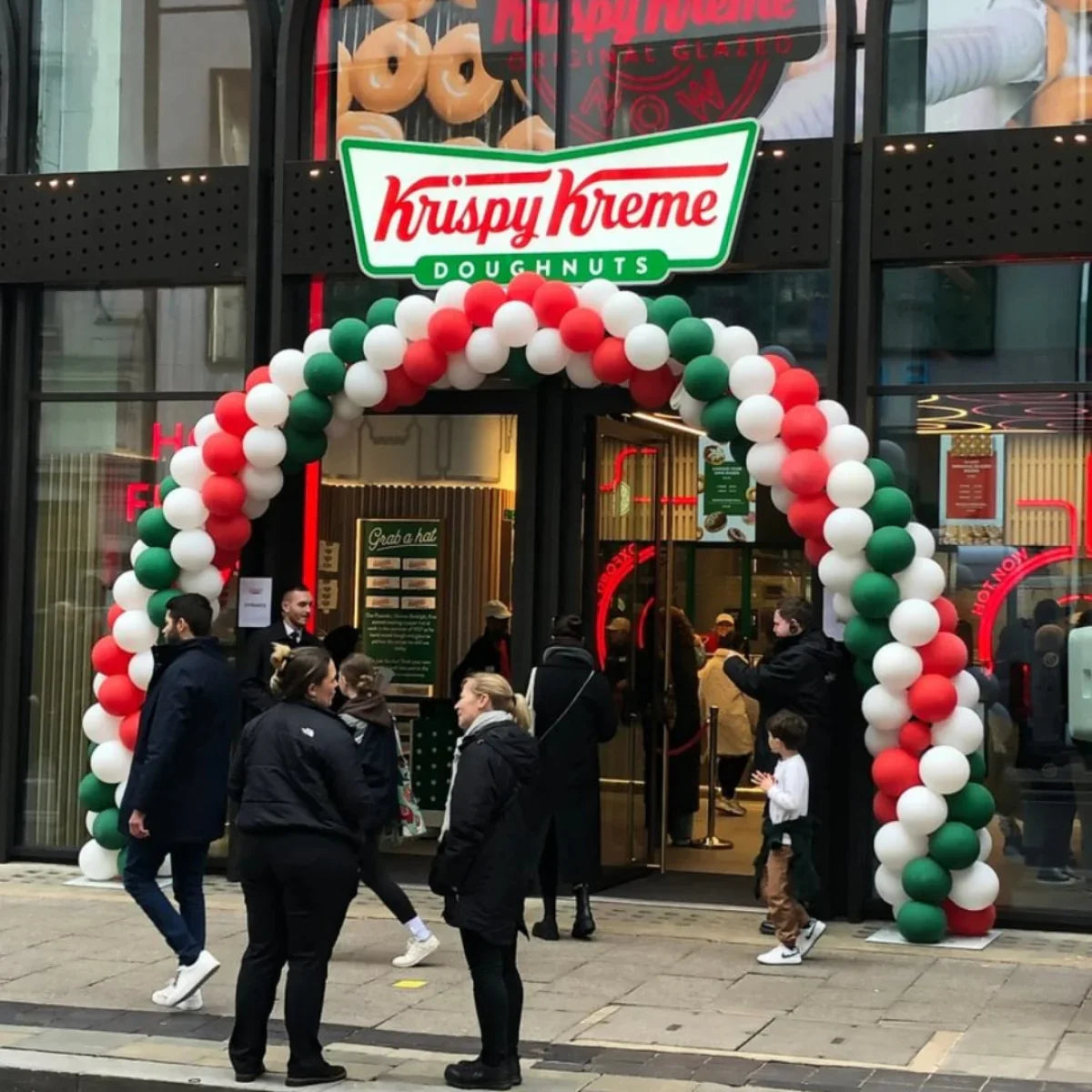 Krispy Kreme store entrance decorated with red, white, and green balloon arch and people walking outside