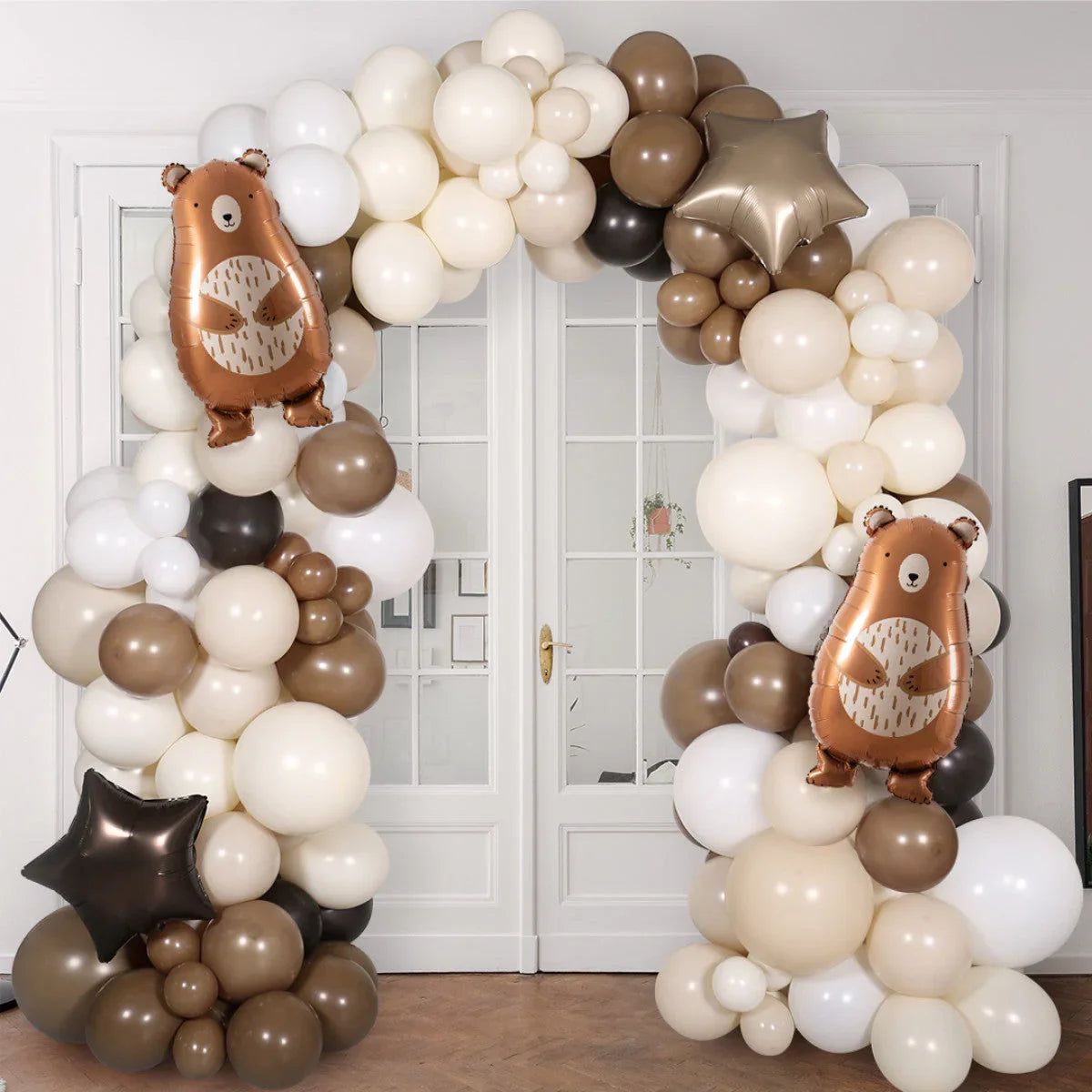 Neutral-tone balloon arch with beige, white, brown balloons and bear-shaped foil balloons in front of glass doors