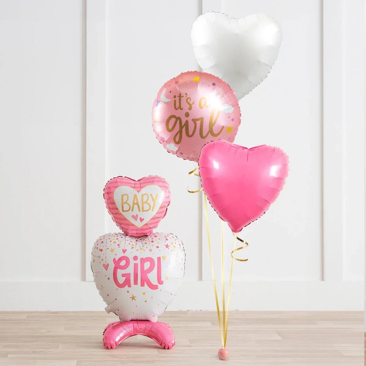 Pink and white heart-shaped and round balloons with 'It's a girl' and 'Baby girl' text on wooden floor