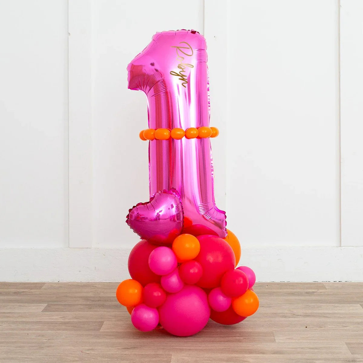 Pink foil balloon shaped as number 1 with orange and pink round balloons decoration on wood floor against white wall