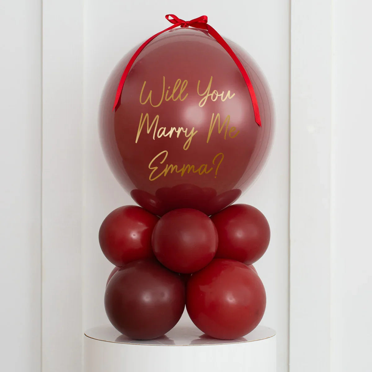 Maroon marriage proposal balloon arrangement with "Will You Marry Me Emma?" text and red ribbon