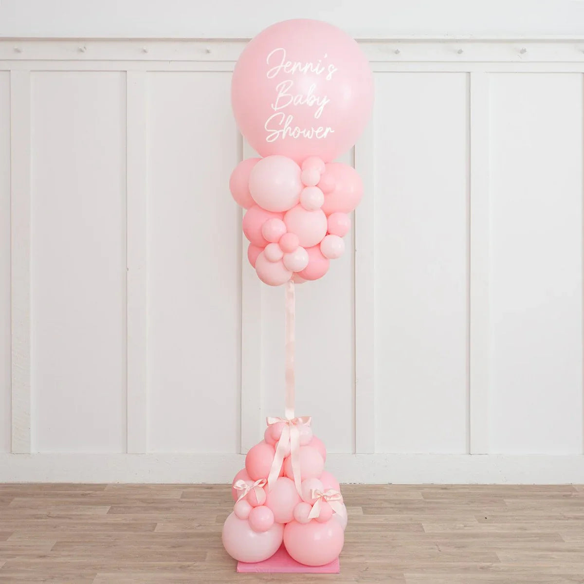Pink balloon pillar decorated for Jenni's baby shower on wooden floor with white paneled wall