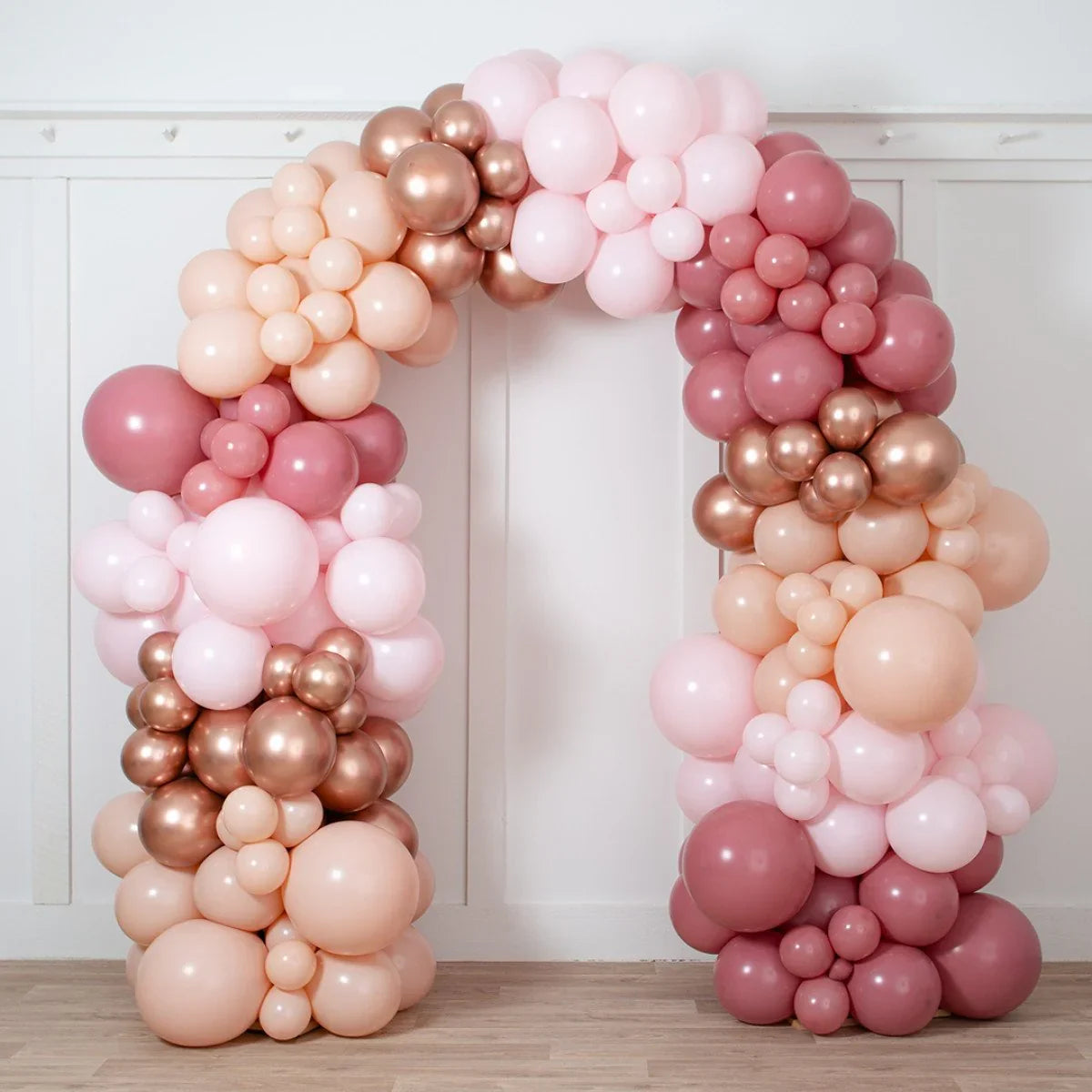 Rose gold balloon arch with shades of pink and peach balloons, set indoors against white paneled wall