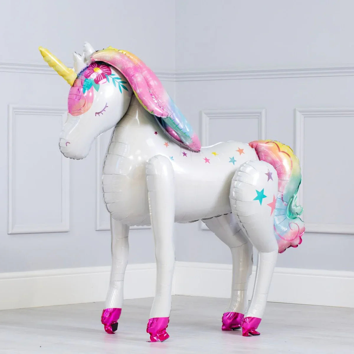 White inflatable unicorn balloon with rainbow mane, tail, yellow horn, and pink hooves standing indoors