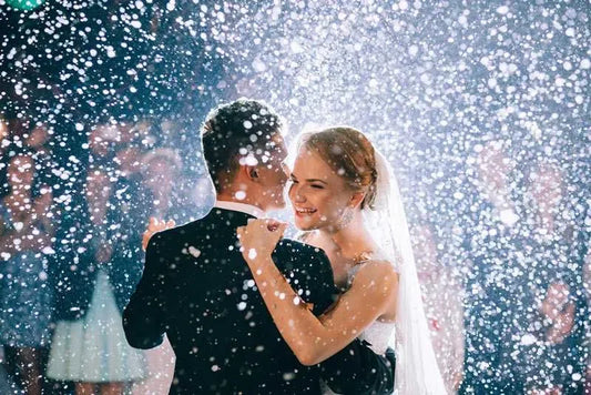 Bride and groom dancing at wedding under falling confetti, surrounded by guests.