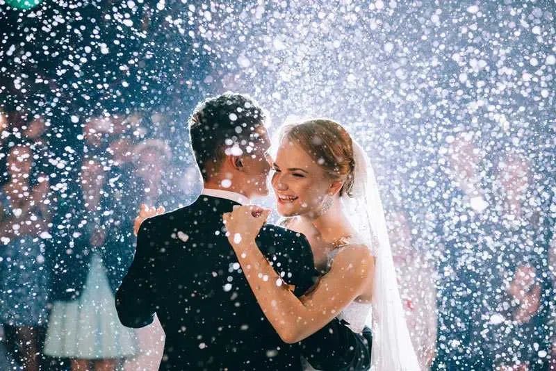 Bride and groom dancing in falling snow at wedding reception with smiling guests in background