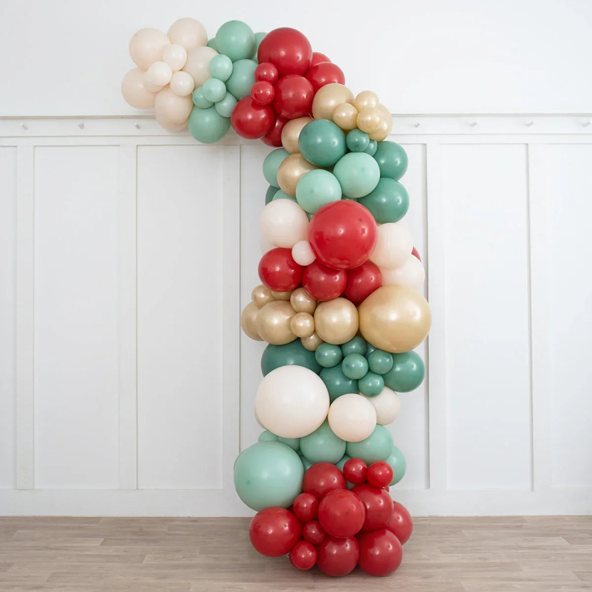 Asymmetric Christmas balloon arch with red, green, cream, and gold balloons in a white room