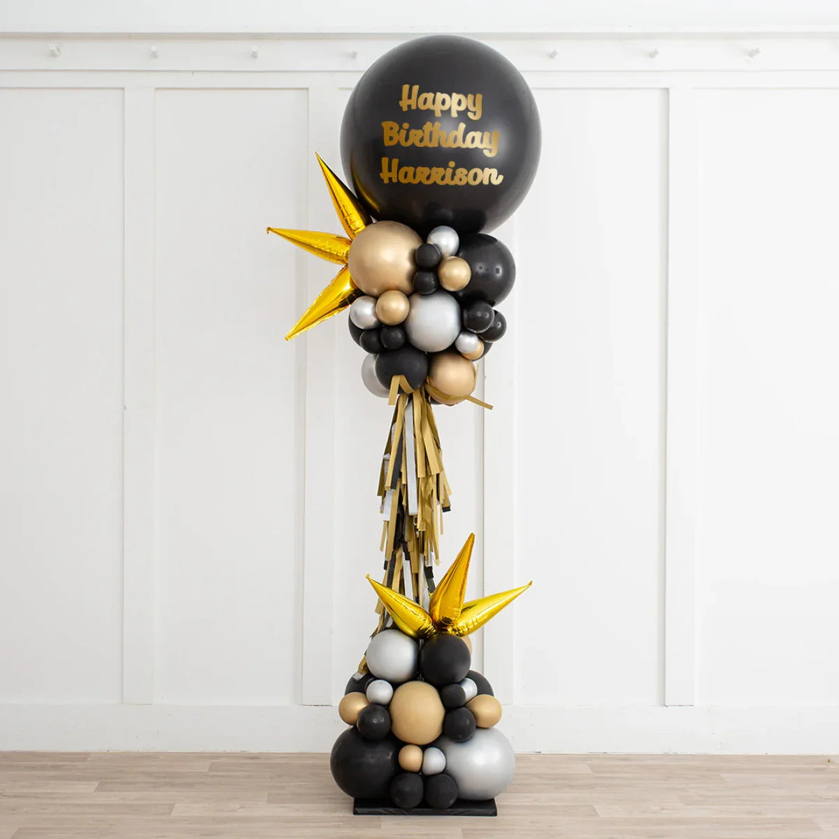 Black, gold, and silver birthday balloon arrangement with 'Happy Birthday Harrison' text in a white room